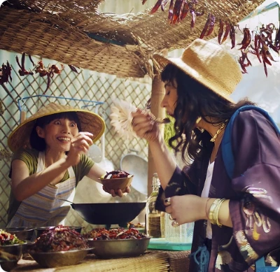 Duas mulheres provando comida de rua sob um mercado com pimentas secas penduradas.